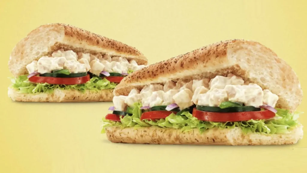 Um banner apresenta dois sanduíches de frango com salada da Subway. Ambos os sanduíches são feitos com pão integral salpicado com sementes, recheados com frango desfiado em molho cremoso, fatias de pepino, fatias de tomate e alface picada. Os sanduíches estão dispostos em um fundo amarelo claro.