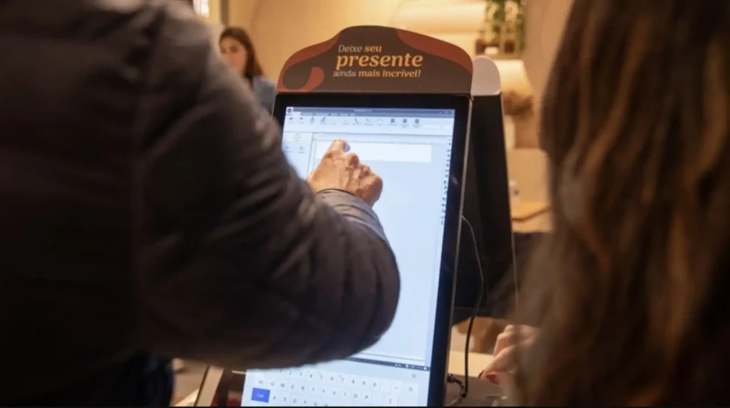 Uma pessoa (parcialmente visível, com um braço e parte das costas expostas) interage com uma tela digital em pé. A tela exibe um software de edição de texto com alguns campos preenchidos. Acima da tela, um adesivo marrom em forma de onda com as palavras "Deixe seu presente ainda mais incrível!" em branco e laranja. Ao fundo, uma segunda pessoa pode ser vista, sentada e parcialmente desfocada, e um ambiente interno com iluminação suave.