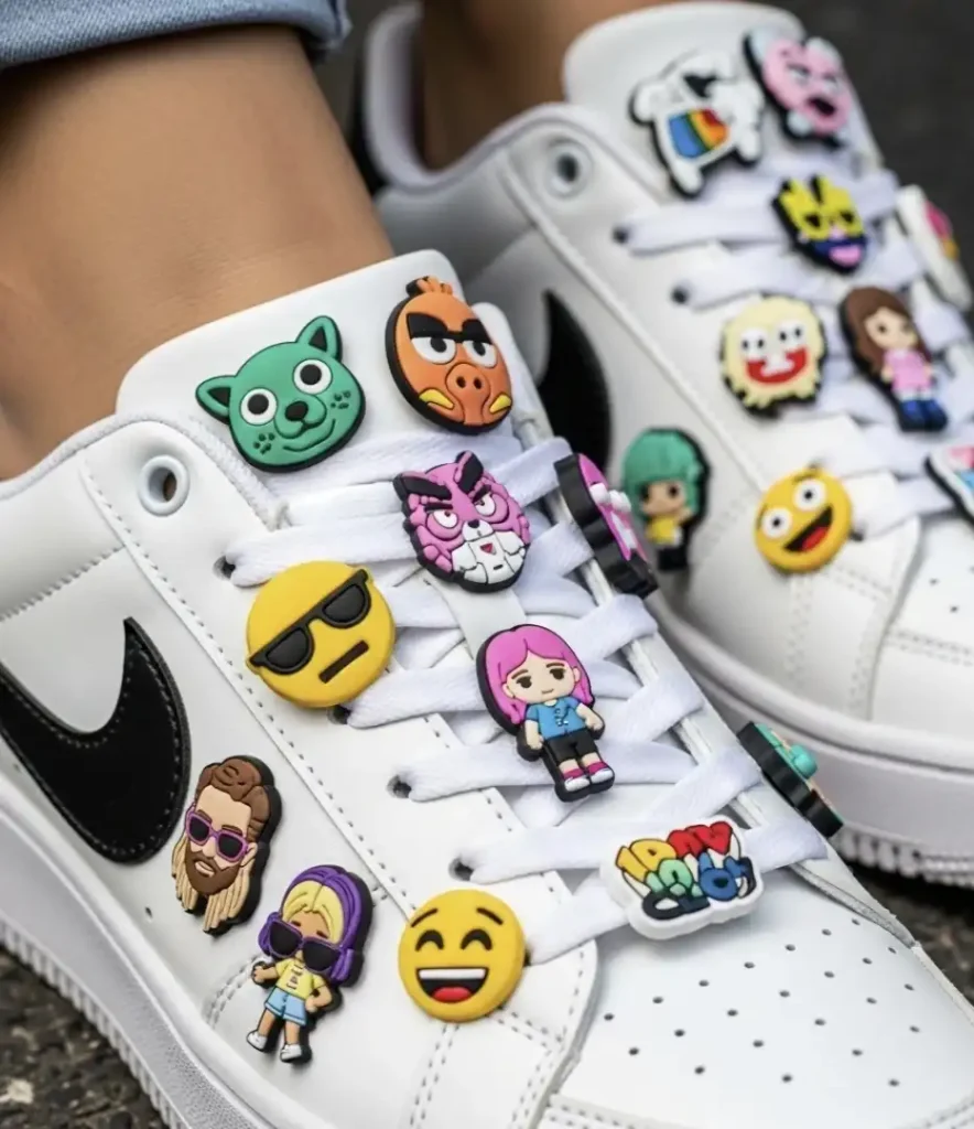 Um par de tênis Nike brancos exibindo uma coleção de charms coloridos e divertidos em seus cadarços. Os charms incluem vários emojis, como um rosto sorridente com óculos de sol e um rosto rindo, bem como personagens de desenhos animados e outras figuras.