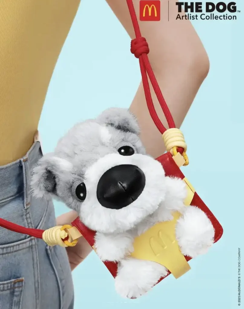 Um close-up de uma pessoa usando uma bolsa pequena, vermelha e amarela, no formato de um cachorro cinza felpudo, com a marca do McDonald's e "The Dog Artlist Collection" no canto superior direito.
