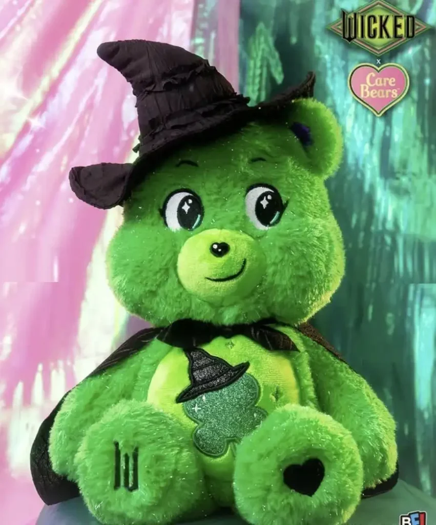 Um Ursinho Carinhoso verde e brilhante, vestido como uma bruxa. Ele usa um chapéu de bruxa preto pontudo e uma capa preta. No centro de sua barriga há uma silhueta preta de um chapéu de bruxa sobre um brilho verde. O logotipo "WICKED" e um logotipo de coração "Care Bears" aparecem na parte superior direita da imagem.