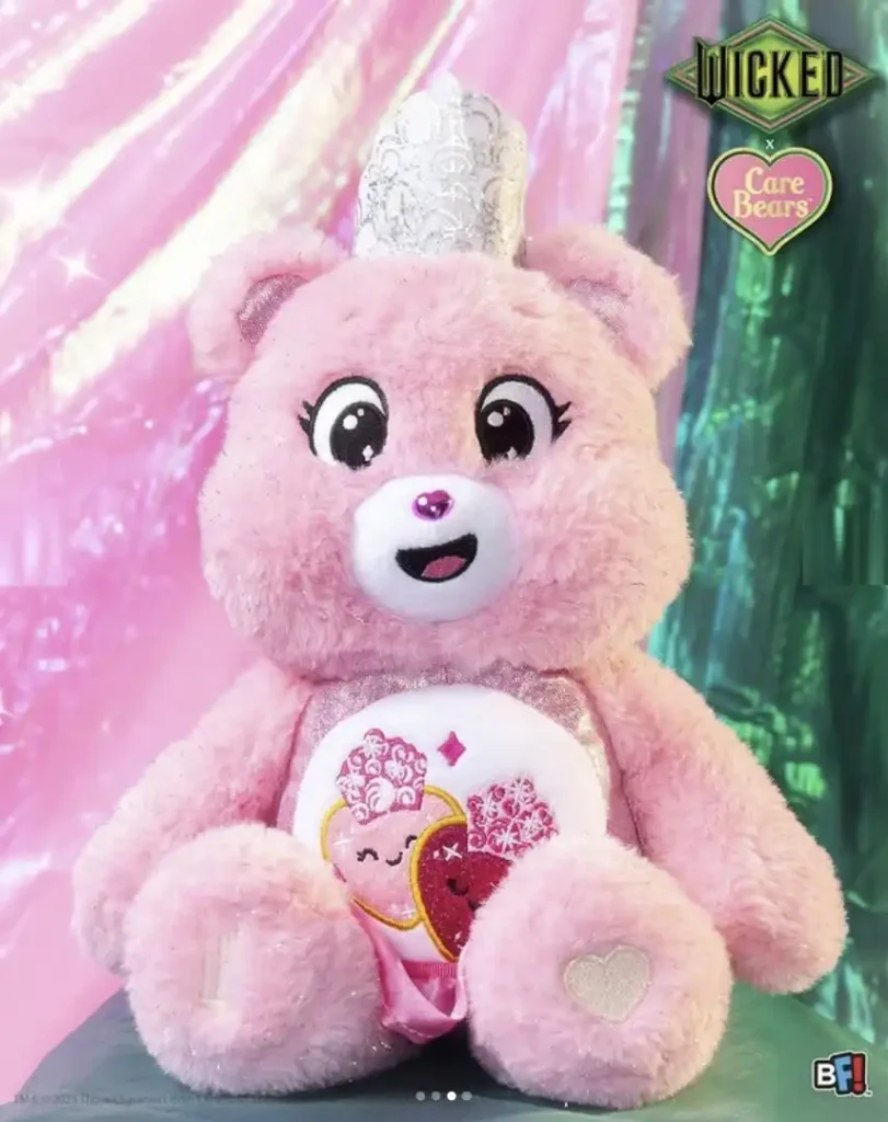 Um Ursinho Carinhoso rosa e brilhante, usando uma coroa prateada e cintilante. Na sua barriga há um design com dois corações sobrepostos, um rosa claro e outro rosa escuro, cada um com uma face sorridente. O logotipo "WICKED" e um logotipo de coração "Care Bears" aparecem na parte superior direita da imagem.