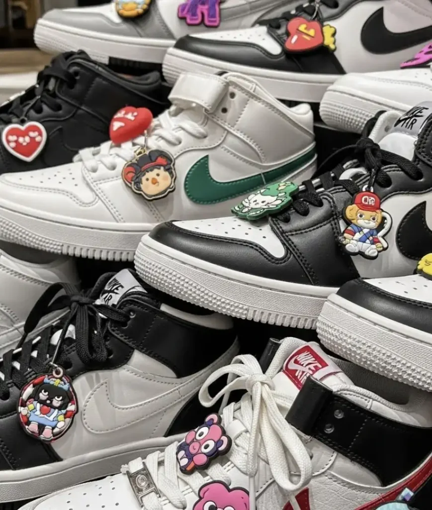 Uma visão de close-up de vários tênis Nike pretos e brancos, incluindo modelos Dunk e Air Force 1. Muitos dos tênis estão enfeitados com pingentes coloridos e de personagens pendurados nos cadarços.