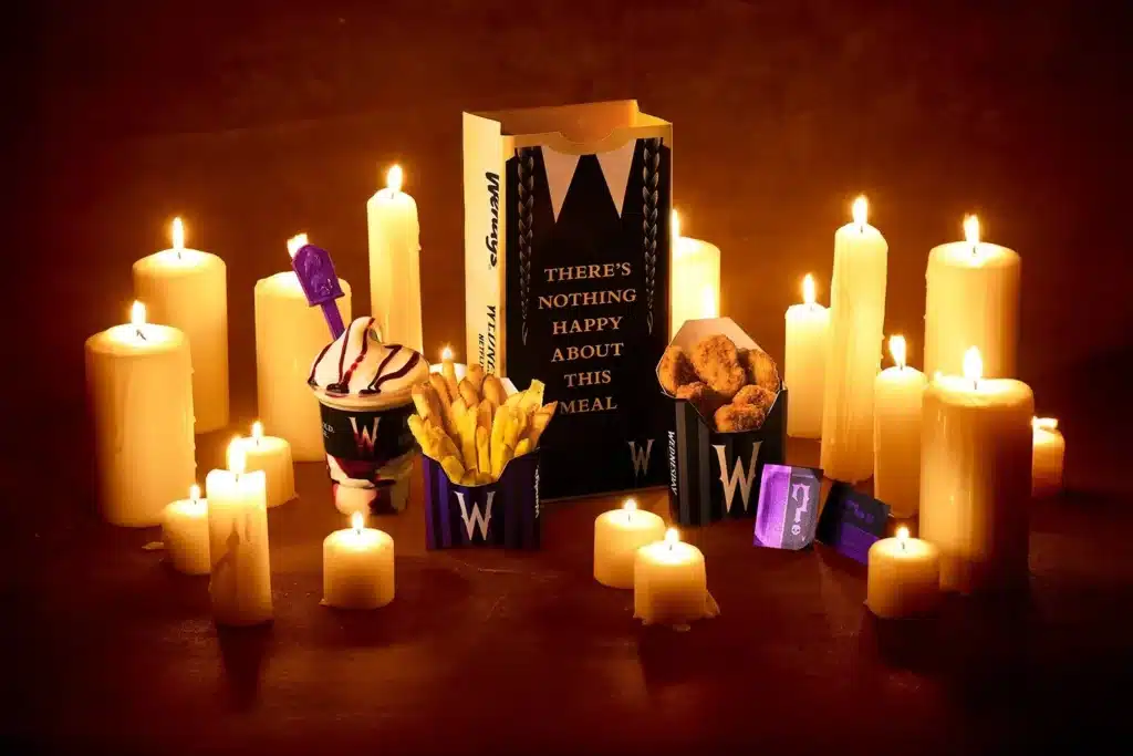 Uma imagem escura e dramática com o tema da Wandinha (Wednesday Addams) mostra um combo de refeição do Wendy's cercado por muitas velas acesas de diferentes tamanhos, criando um brilho quente. No centro, uma caixa alta e preta com o logotipo do Wendy's e a frase "There's nothing happy about this meal" (Não há nada de feliz nesta refeição) serve como pano de fundo. À sua frente, estão os itens do combo: um copo com sorvete de chocolate e creme, batatas fritas em um pacote roxo e preto com a letra "W" estilizada, e um pacote de nuggets de frango semelhante. Todos os itens de comida têm embalagens com o tema da Wandinha. A iluminação das velas realça as sombras e cria uma atmosfera misteriosa.