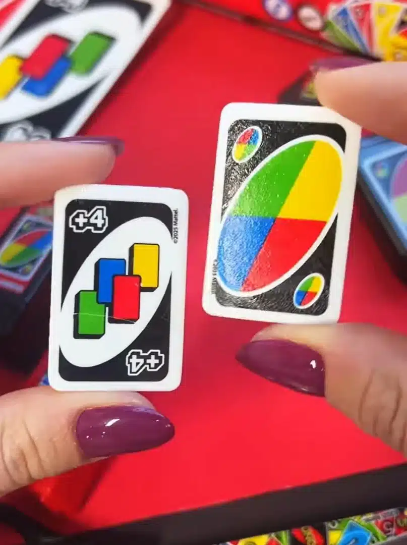 Duas cartas de borracha UNO em miniatura, uma 'Wild' e uma '+4', sendo seguradas por uma pessoa sobre um fundo vermelho.