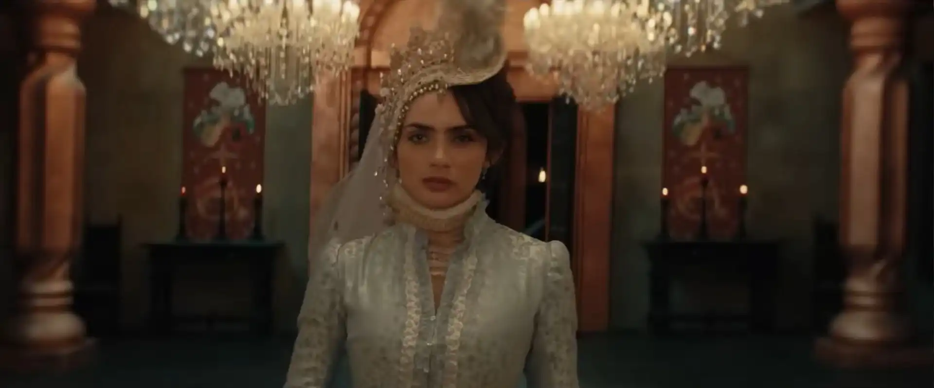 Uma mulher de cabelos castanhos escuros e olhos azuis, com uma expressão séria, está em pé em um salão opulento.