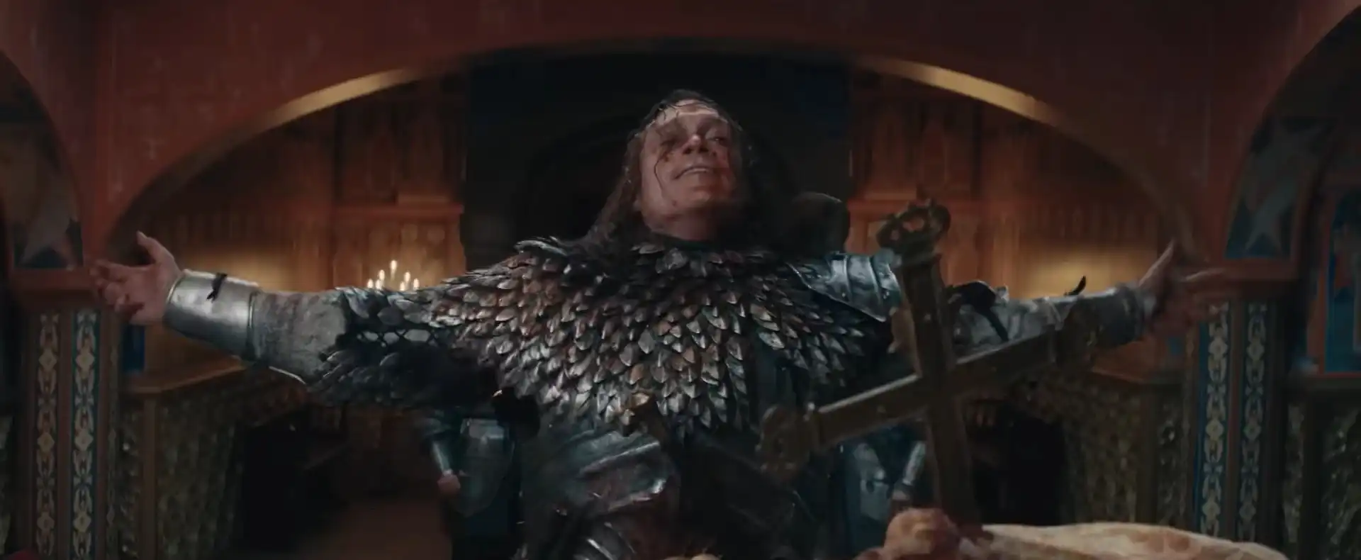 Um homem de meia-idade, com cabelos castanhos longos e rosto machucado e ensanguentado, veste uma armadura escura com detalhes que lembram escamas e braços de metal.