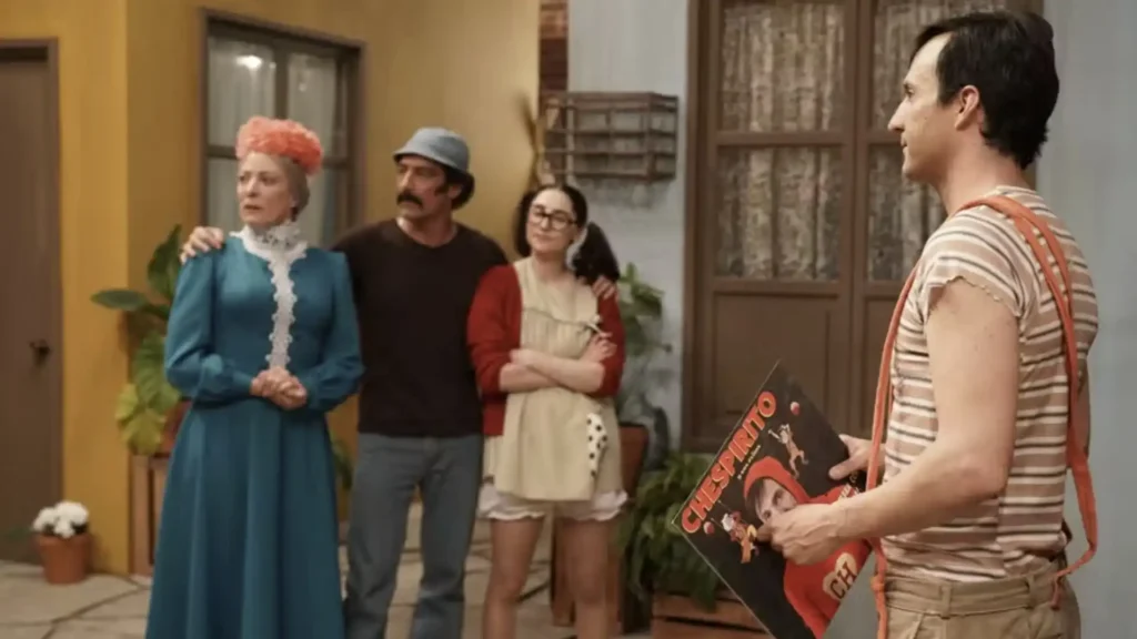 Uma imagem mostra quatro atores interpretando personagens da famosa série "Chaves", criada por Chespirito. A cena se passa no pátio da vila. Da esquerda para a direita, estão uma mulher mais velha de cabelos cor-de-rosa e vestido azul, um homem com bigode e chapéu, uma jovem de óculos e avental, e um homem à direita, de costas, vestindo uma camiseta listrada e segurando um objeto retangular com a imagem do Chaves.