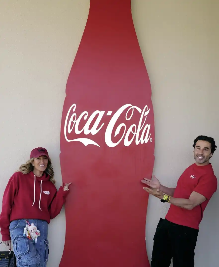Um homem e uma mulher, ambos vestindo roupas vermelhas, posam ao lado de um grande banner vermelho da Coca-Cola.