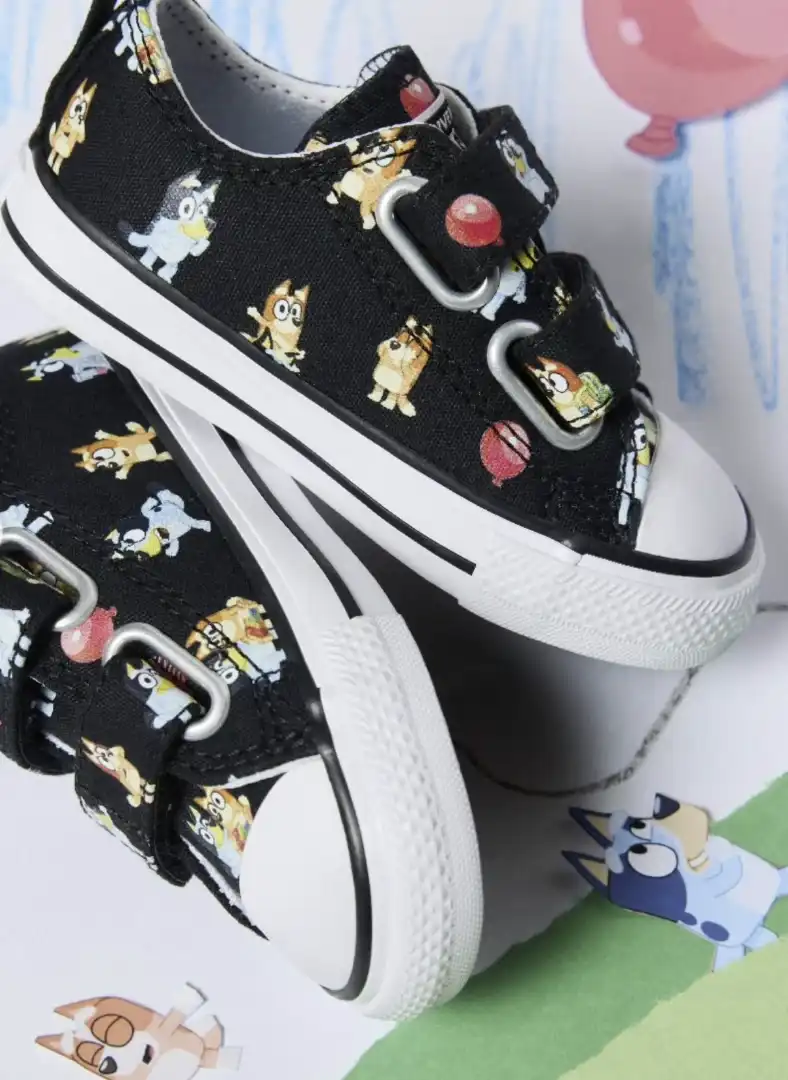 Um par de tênis Converse preto, de cano baixo, com estampa dos personagens da animação Bluey. Os cadarços são substituídos por fechos de velcro, e a sola de borracha branca tem um detalhe em linha preta. Ao fundo, há desenhos dos personagens Bluey e Bingo.