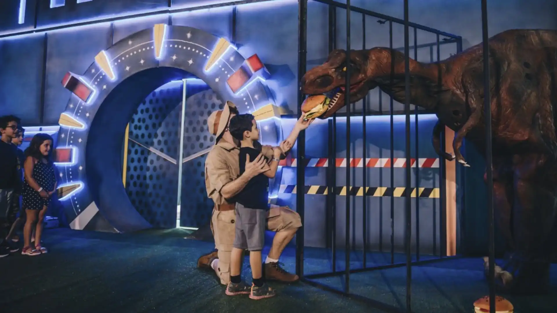 Homem vestido como guarda florestal alimenta um T-Rex de brinquedo através de uma jaula, enquanto crianças observam em um cenário temático com portão futurista iluminado.