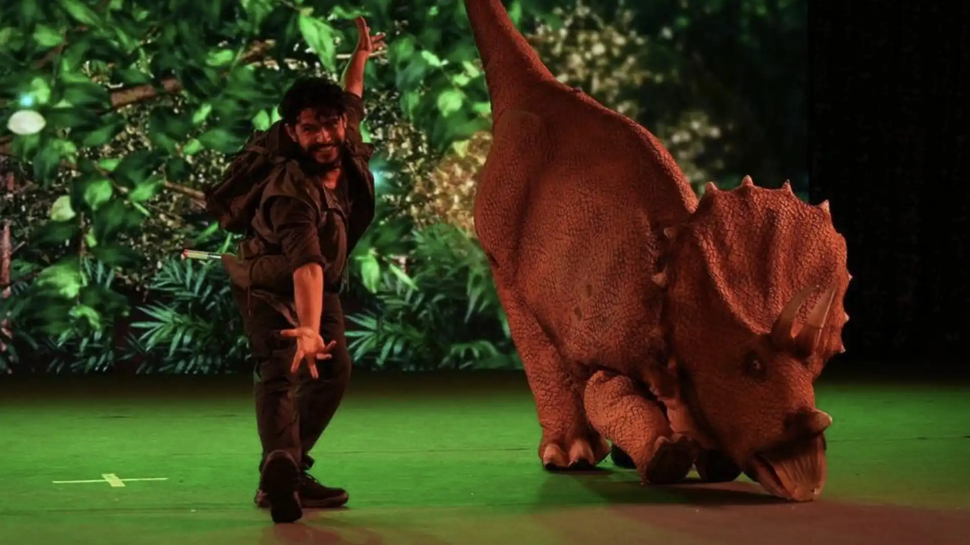 Homem em traje de explorador acena com a mão, em pose descontraída, ao lado de uma réplica de triceratops em um palco com um telão de floresta ao fundo.