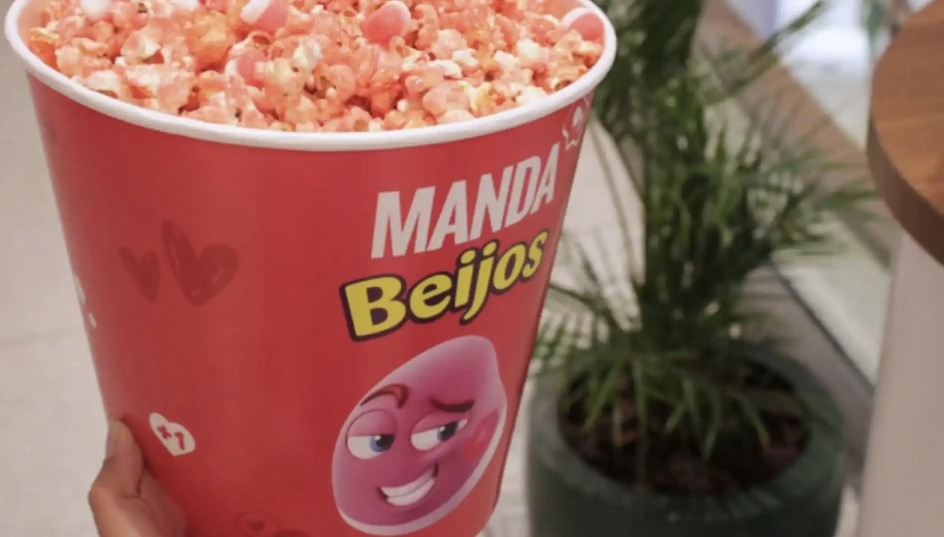Pessoa segurando um balde de pipoca grande do Cinemark, com a estampa "Manda Beijos" e pipoca sabor Fini, de cor avermelhada e com doces brancos.