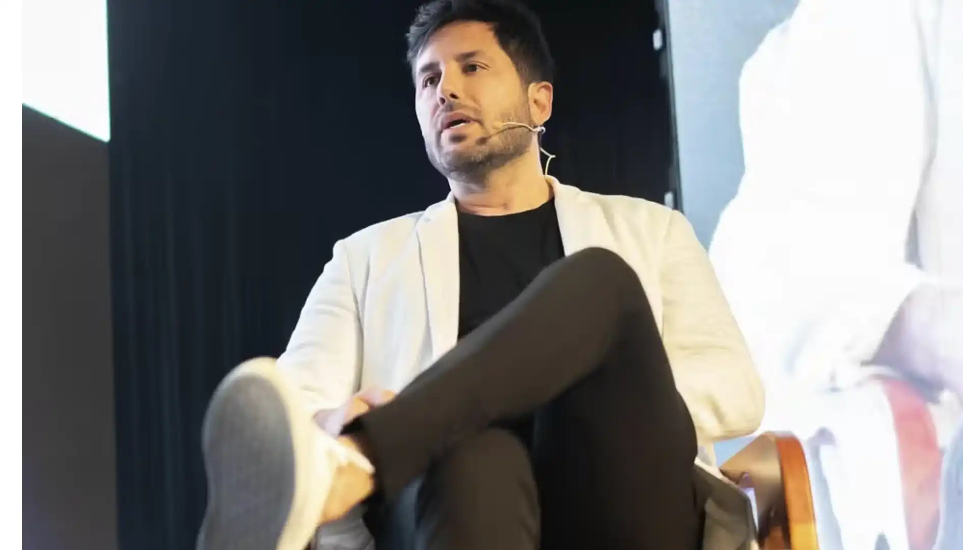 Um homem com barba, vestindo blazer branco e camiseta preta, sentado e falando em um microfone de lapela.