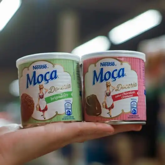 Duas latas de doces de colher da linha Moça Doceria, sabor Brigadeiro e Sabor Chocolate Cremoso, sendo seguradas por uma mão.