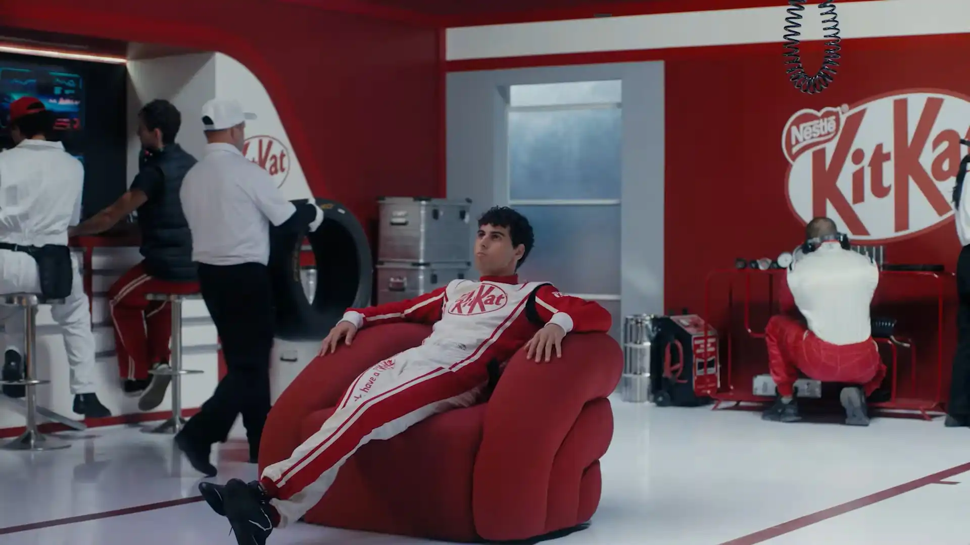 Um homem em um macacão de corrida KitKat relaxa em uma poltrona em formato de KitKat em uma sala temática de corrida, com outras pessoas ao fundo.