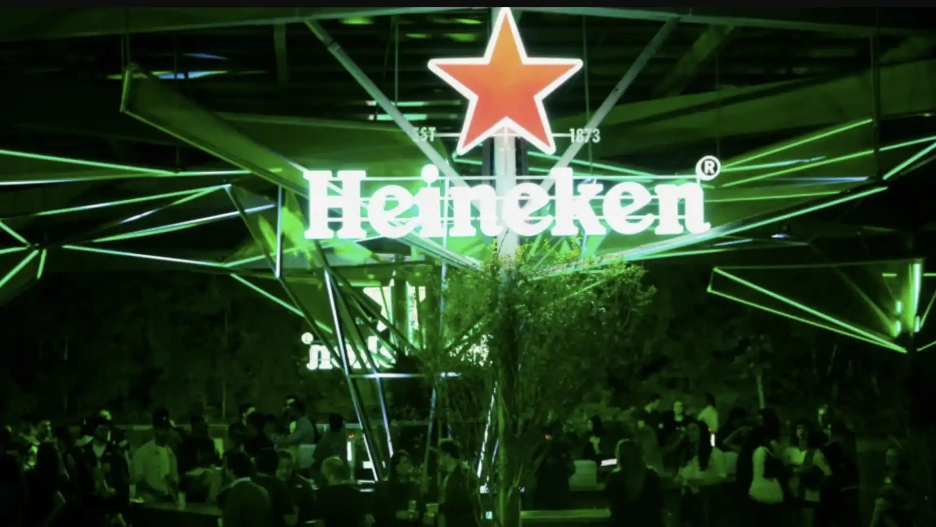 Imagem de uma balsa-bar da Heineken flutuando em um rio, com a silhueta de uma cidade ao fundo.