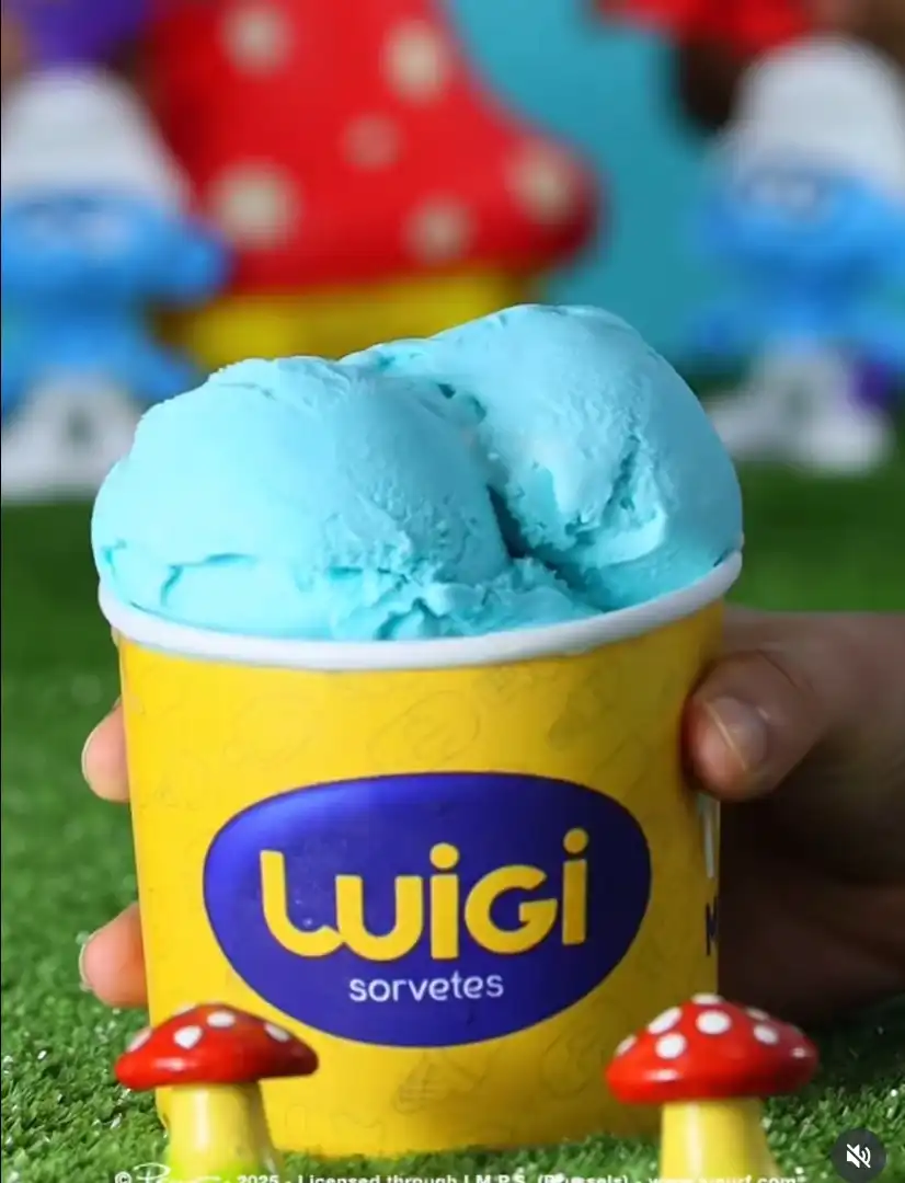 Uma mão segurando um copo amarelo da marca Luigi Sorvetes, contendo duas bolas de sorvete azul-claro que lembram o sabor "Smurfs". O fundo mostra bonecos dos Smurfs e cogumelos, recriando o ambiente da vila dos personagens.