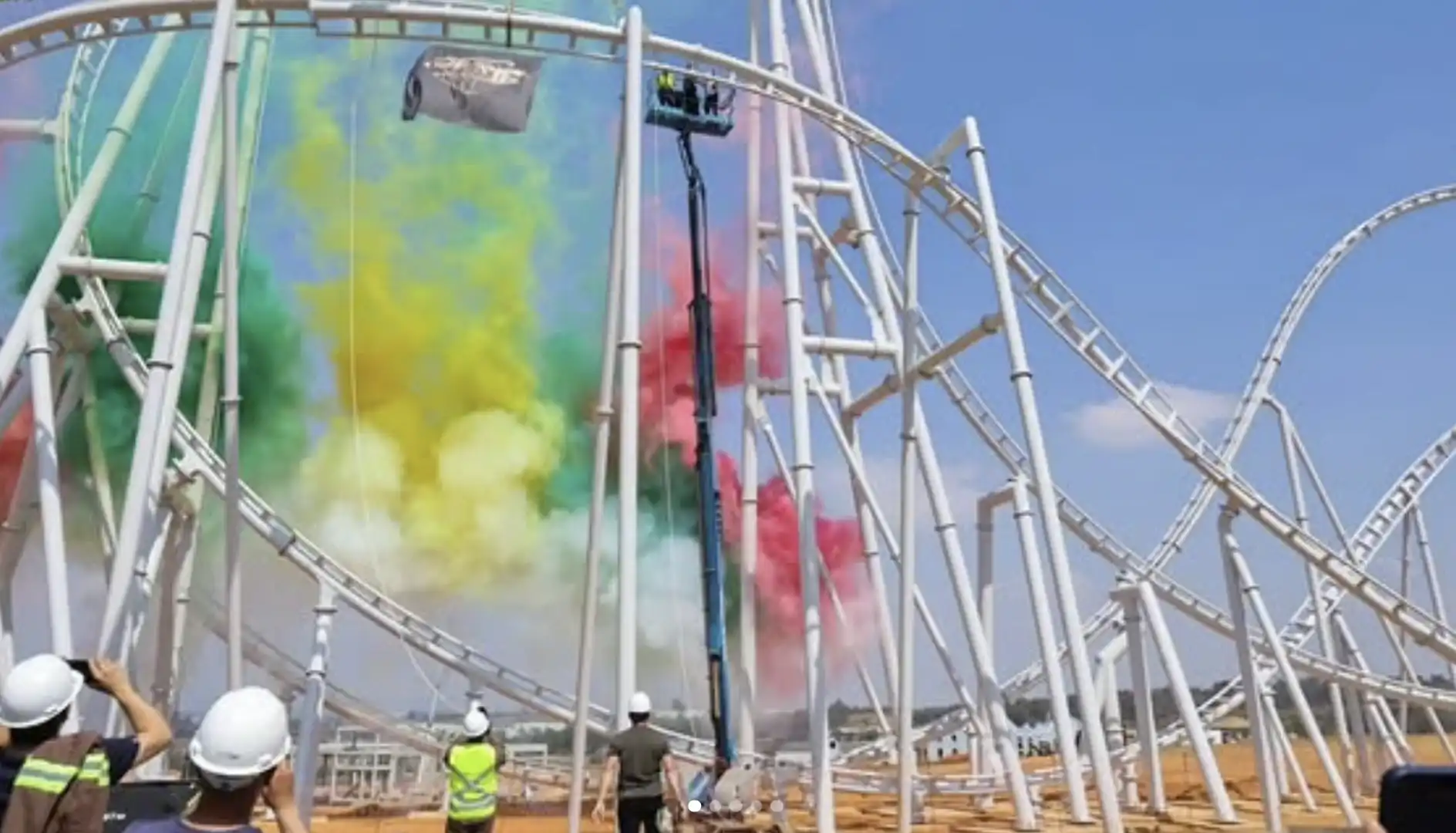 Imagem de uma montanha-russa branca no Cacau Park com jatos de fumaça colorida verde, amarela e vermelha durante uma vistoria ou evento. Pessoas usando capacetes de segurança observam.