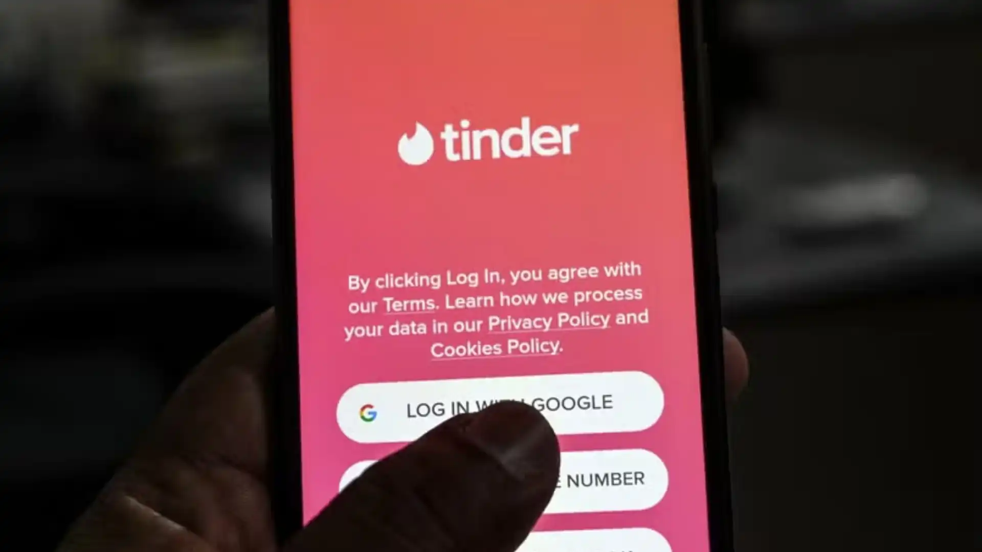 A mão de uma pessoa é mostrada tocando o botão "Fazer login com o Google" na tela de login do aplicativo Tinder.