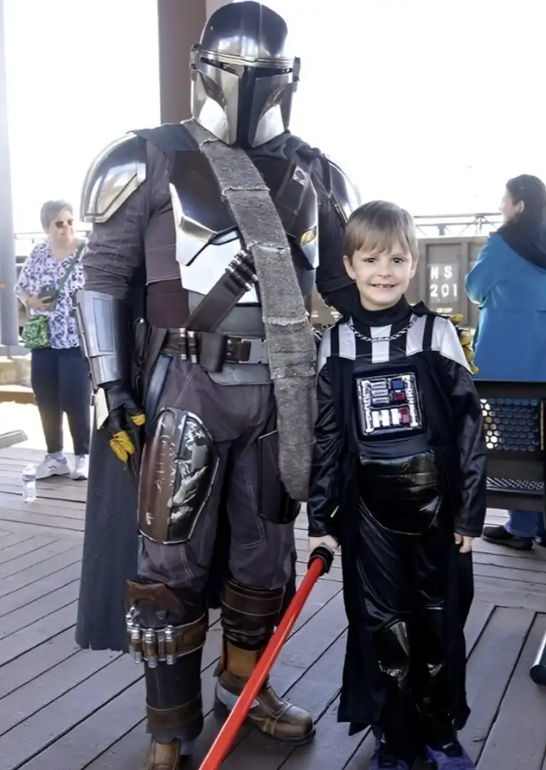 Uma criança vestida de Darth Vader, segurando um sabre de luz vermelho, posa ao lado de uma pessoa com a armadura completa do Mandaloriano.