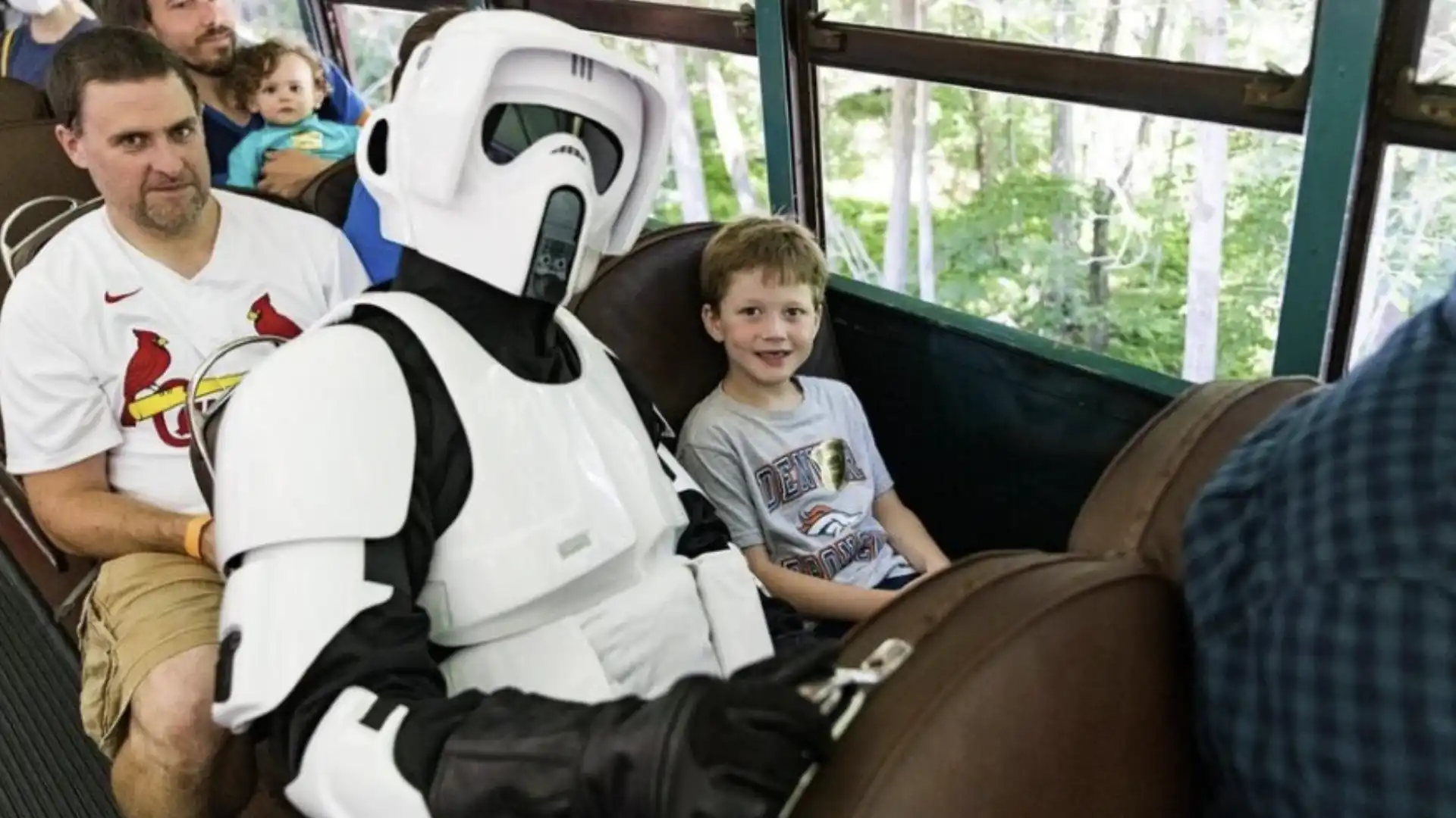 Um Scout Trooper de Star Wars está sentado em um trem com um garoto sorrindo e um homem barbudo com um bebê no colo.