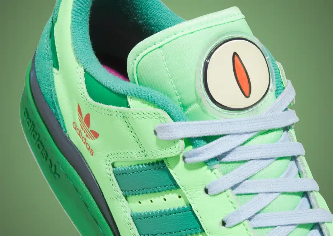Um close-up de um tênis Adidas Forum Low "The Simpsons Treehouse of Horror" verde. A imagem destaca a língua do tênis que possui um olho grande e circular com uma íris laranja, cadarços azuis claros, as três listras verdes escuras da Adidas e o logotipo laranja da Adidas na lateral.