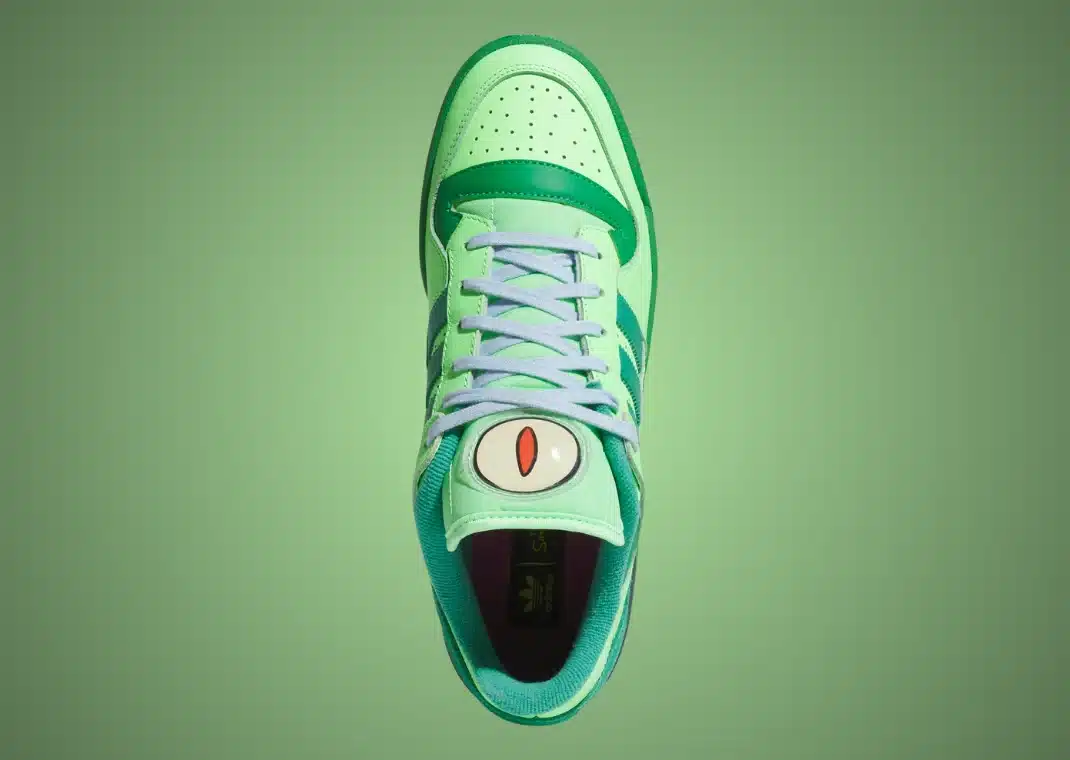 Uma vista de cima de um tênis Adidas Forum Low "The Simpsons Treehouse of Horror" com o desenho do olho do personagem Kodos na língua. O tênis é predominantemente verde, com três listras em verde-escuro, cadarços azuis e um interior roxo.