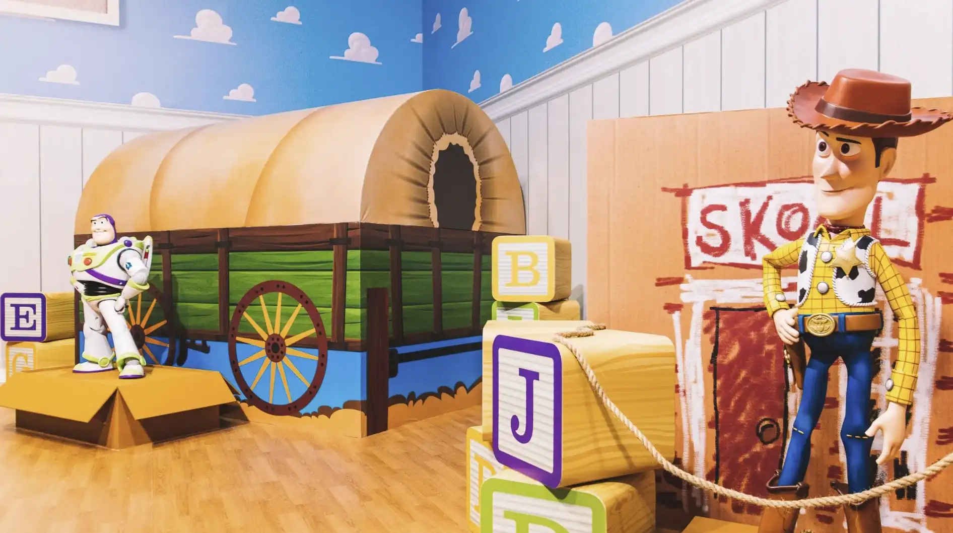 A imagem mostra Woody e Buzz Lightyear de Toy Story em um quarto com paredes de céu azul e nuvens, ao lado de uma carroça de brinquedo. Blocos de letras estão no chão.
