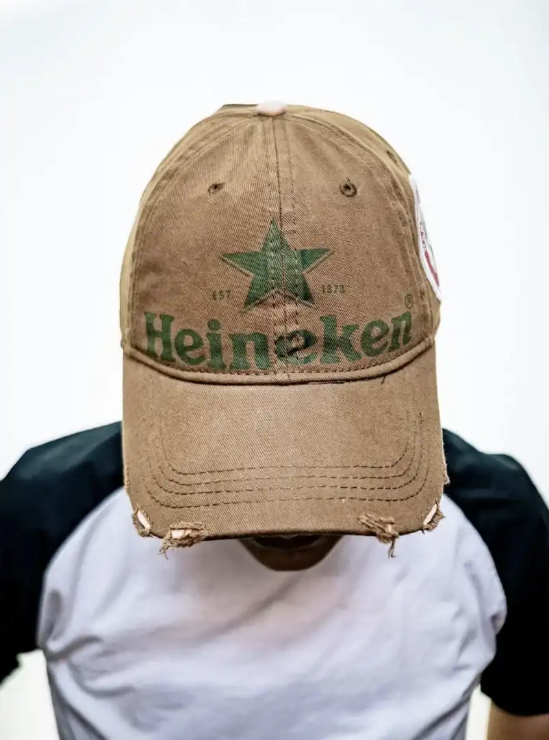 Pessoa usando um boné marrom e desgastado da Heineken, que cobre o rosto, contra um fundo branco.