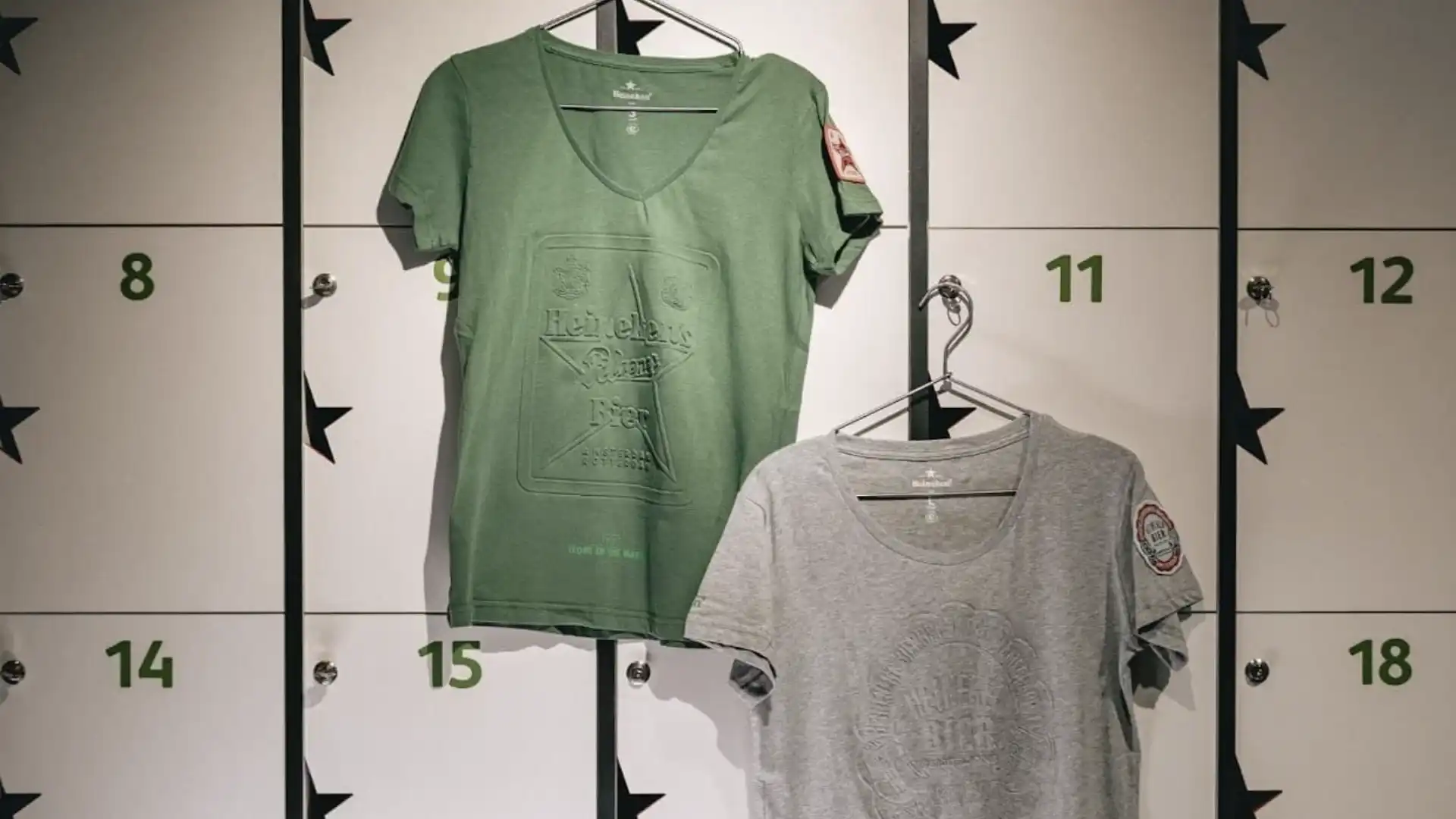 Duas camisetas, uma verde e uma cinza, penduradas em armários numerados com estrelas, em um provador.