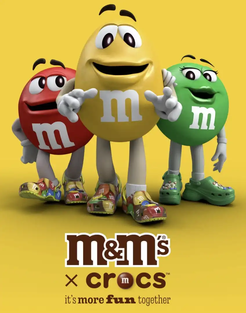 Uma imagem de três personagens de M&M's, o vermelho, o amarelo e a verde, calçando Crocs com a estampa de M&M's. O logo da parceria "M&M's x Crocs" está na parte inferior da imagem.