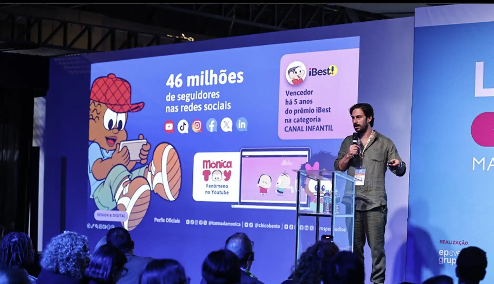 Foto de um homem em pé no palco com um microfone, apresentando para uma plateia em um evento escuro.
