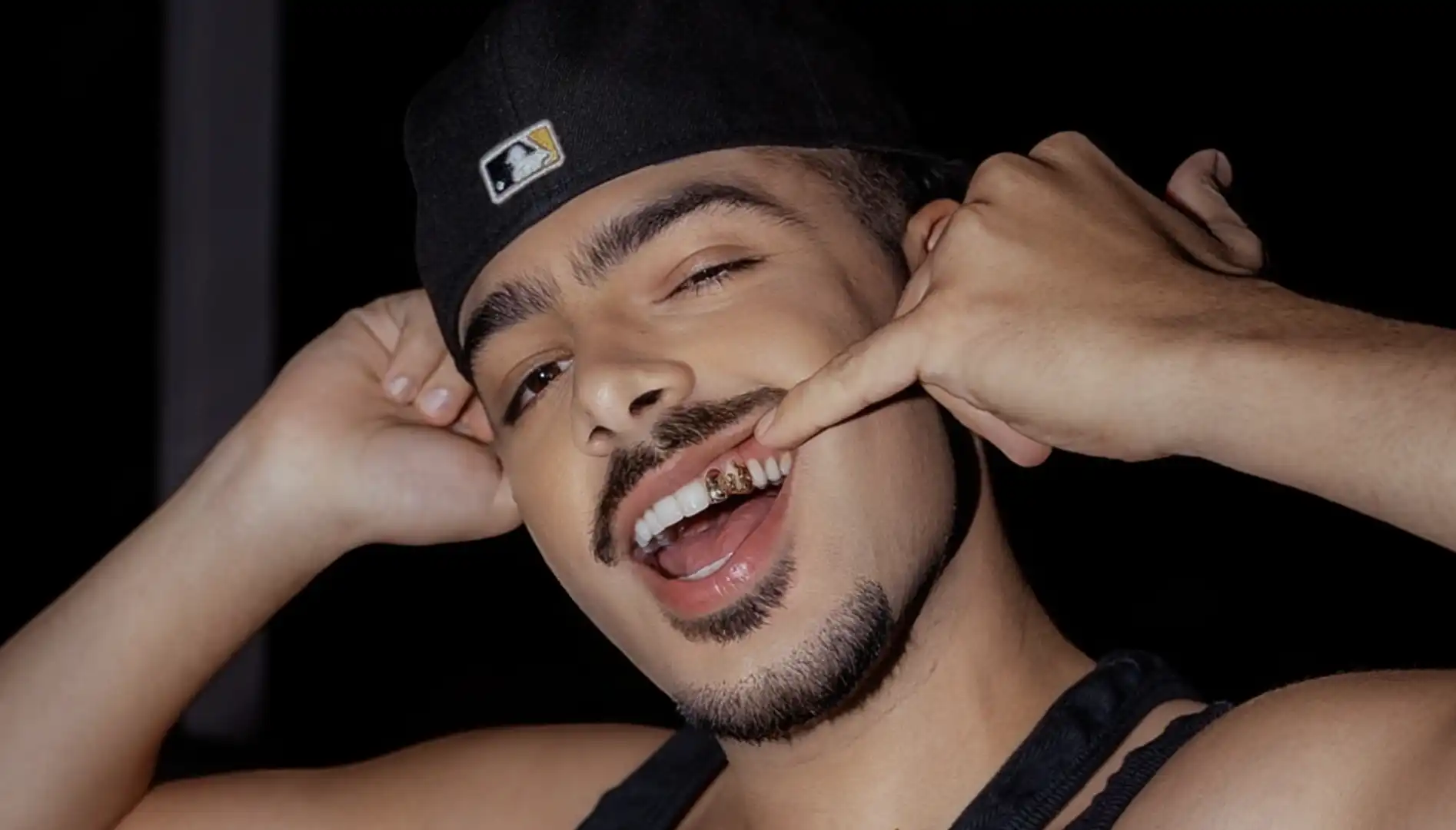 O DJ e produtor musical Pedro Sampaio com um sorriso, usando grillz dourados nos dentes e um boné preto.
