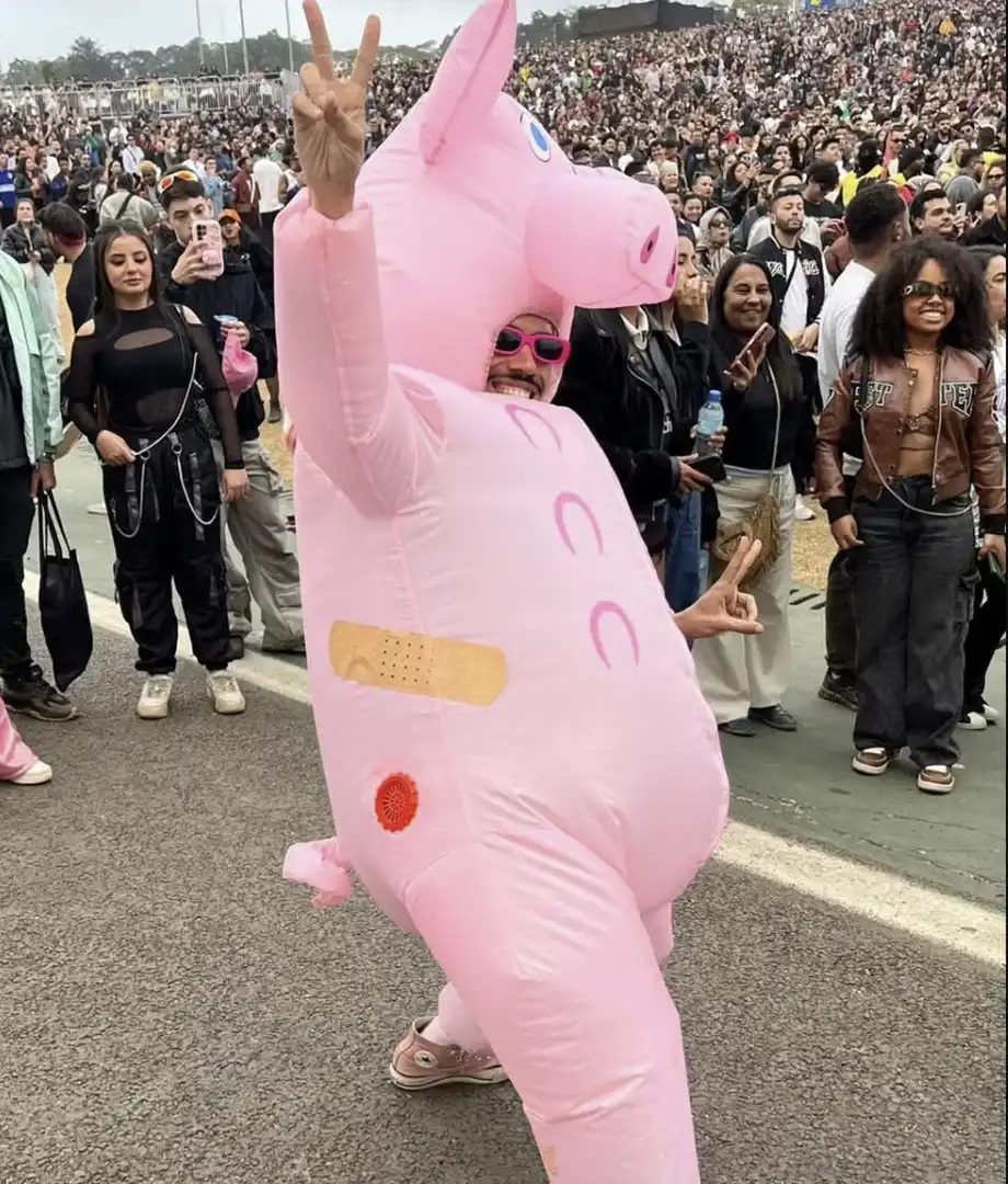 Homem posando para uma foto em um festival de música The Town, usando uma fantasia inflável de porquinho rosa, com um curativo nas costas e óculos de sol, enquanto acena com o sinal de paz.