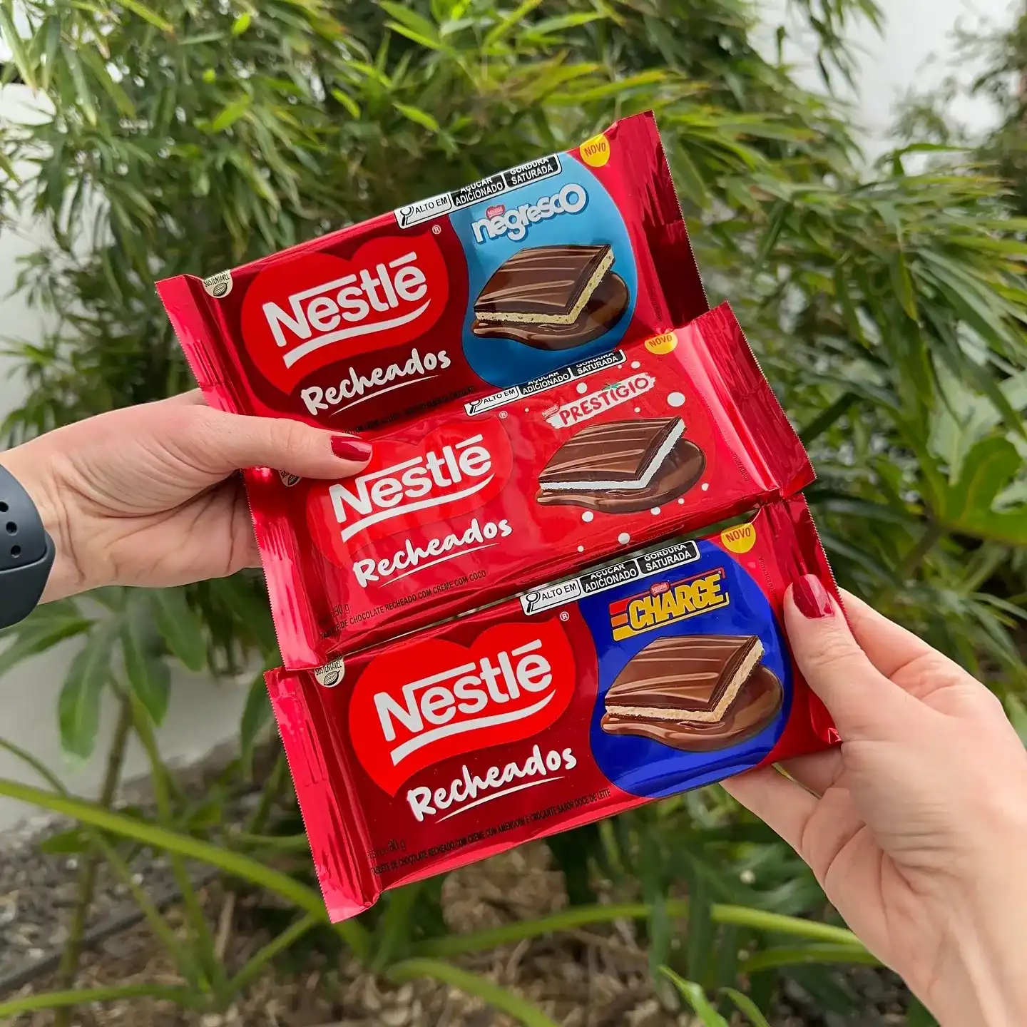 Três embalagens vermelhas de biscoito recheado Nestlé, com os sabores Negresco (azul), Prestígio (vermelho) e Charge (azul), sendo seguradas por duas mãos em frente a um fundo de folhagens verdes.
