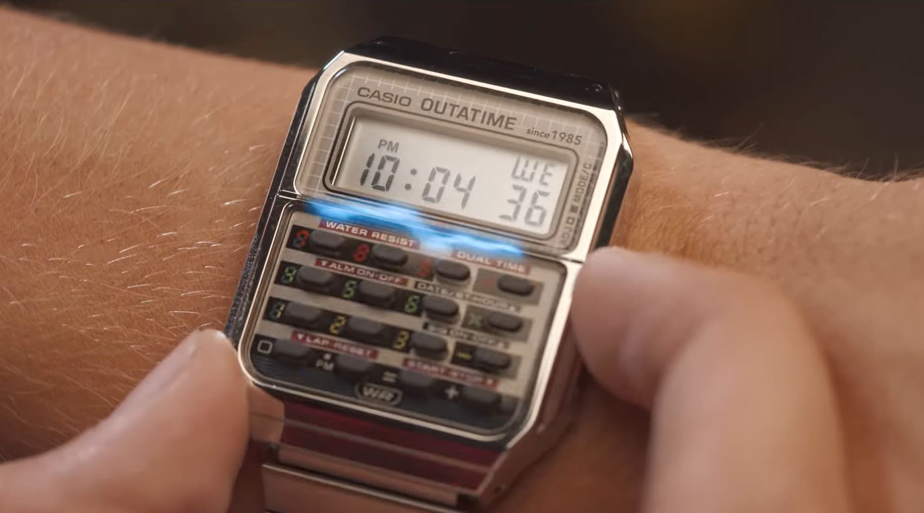 Close-up de um relógio Casio digital com calculadora no pulso, com o texto "CASIO OUTATIME" e um efeito azul neon na tela, fazendo referência a "De Volta Para o Futuro".