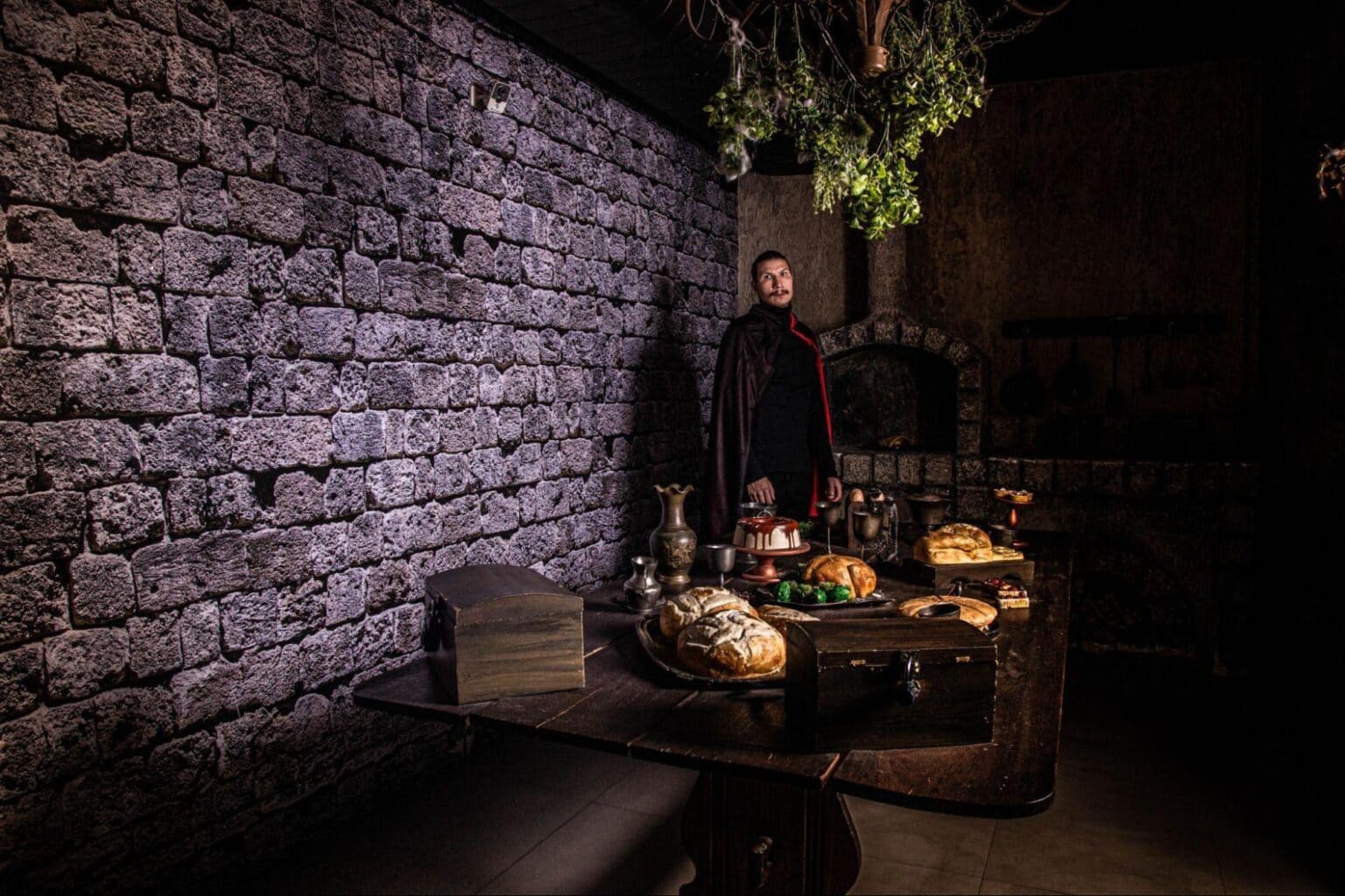 Homem em fantasia de Drácula em uma sala com paredes de pedra, perto de uma mesa rústica com comida e objetos.