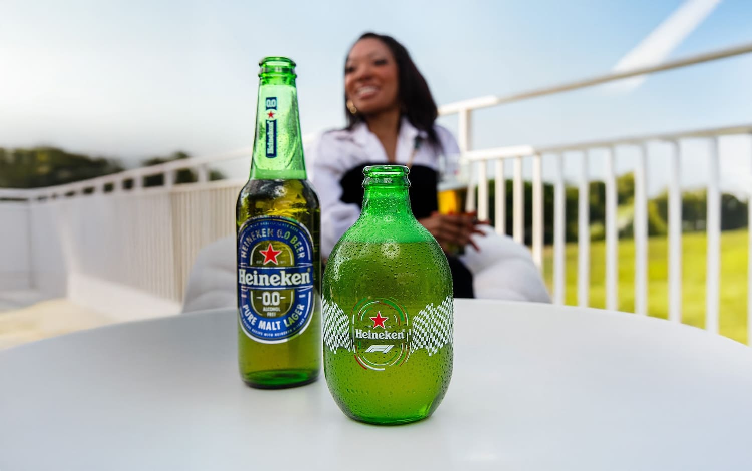Duas garrafas de Heineken sobre uma mesa, uma delas com design especial da Fórmula 1, com uma mulher sorrindo ao fundo.