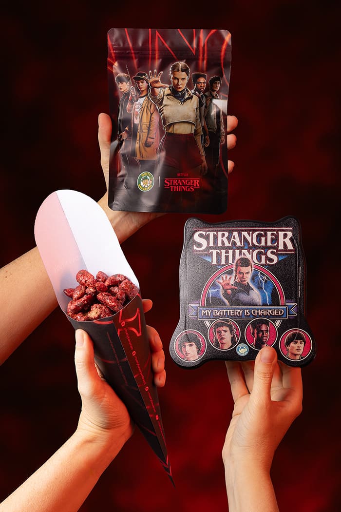 Duas mãos seguram um pacote de nozes temático de Stranger Things com a Eleven e outros personagens, e a outra mão segura um cone de papel preto e vermelho cheio de nozes caramelizadas, ao lado de uma lata de colecionador em forma de cabeça de gato com o logo da série.