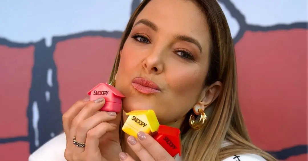 Ticiane Pinheiro fazendo bico e segurando três balms colecionáveis em formato de casinhas do Snoopy, nas cores rosa, amarelo e vermelho.