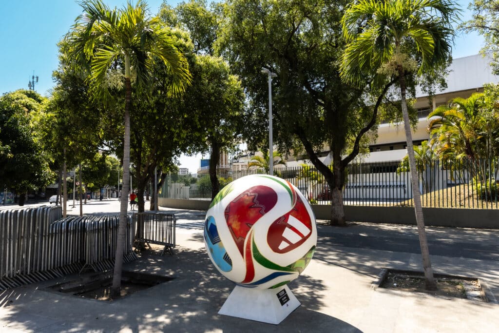 Uma esfera gigante, colorida e decorada, inspirada na bola oficial da Copa do Mundo, está exposta sobre um pedestal branco em uma calçada ensolarada. A bola tem um design vibrante em branco, vermelho, azul e verde. Ao redor, há palmeiras, outras árvores frondosas e grades de contenção de metal, com uma rua e edifícios ao fundo.