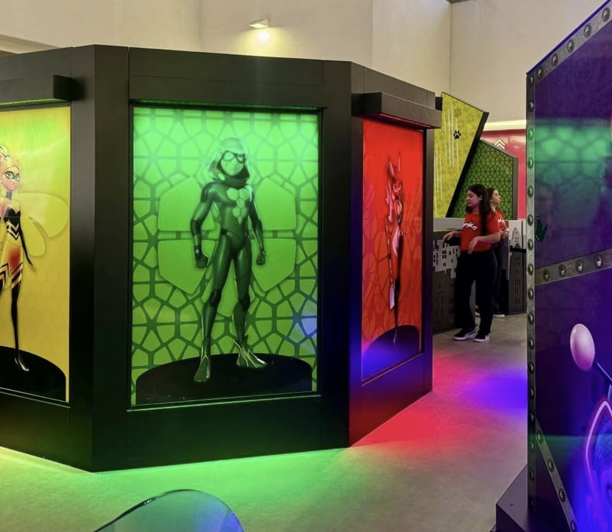 Estrutura hexagonal escura em uma ativação de shopping com painéis iluminados de Queen Bee, Cat Noir e Rena Rouge.