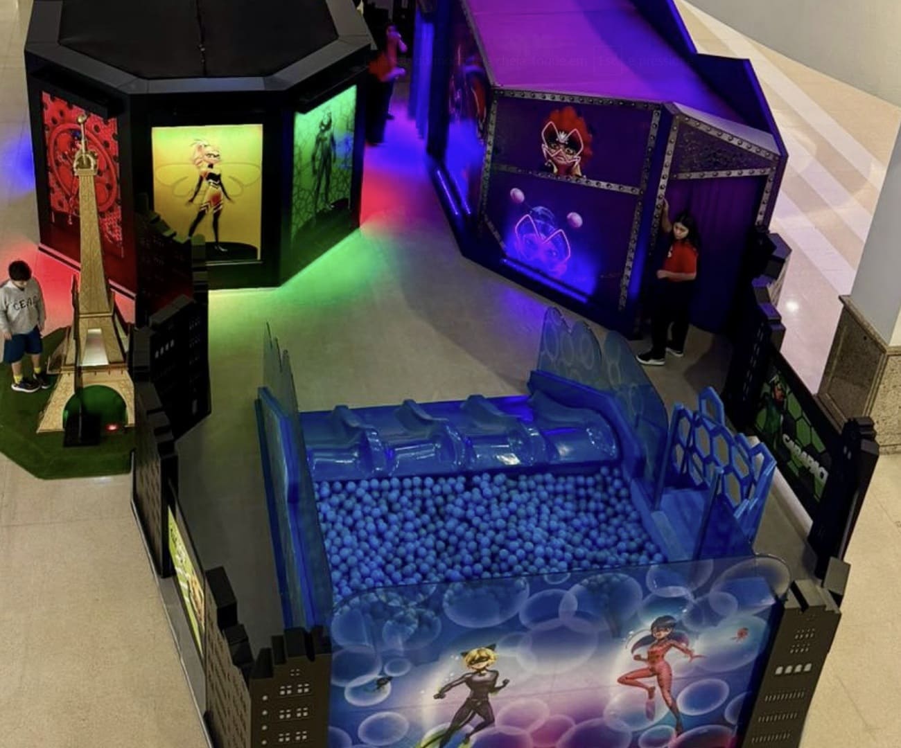 Área de lazer infantil temática de Miraculous: As Aventuras de Ladybug. O espaço inclui uma piscina de bolinhas azuis e estruturas pretas decoradas com os personagens e a Torre Eiffel.