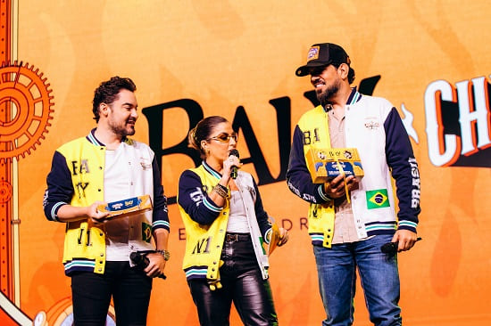 Fernando, uma mulher ao microfone, e Sorocaba no palco em evento da Baly Energy Drink e Churrasco On Fire. Os três vestem jaquetas amarelas, brancas e azuis.