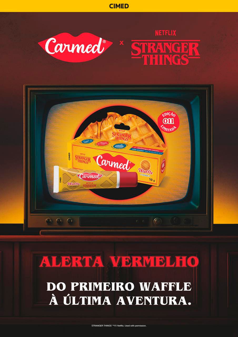 Um Carmed labial de edição especial 'Stranger Things' sabor Waffle, de embalagem amarela e vermelha, em primeiro plano sobre uma superfície de madeira, com sua caixa que imita a textura de um waffle no fundo.