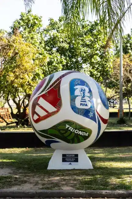 Uma bola gigante e colorida, representando o modelo "Trionda" da Copa do Mundo, está exposta em um pedestal branco sobre um gramado, com árvores e vegetação ao fundo sob luz natural. A bola apresenta um design com painéis em branco, vermelho, verde, azul e dourado, e exibe o emblema da FIFA World Cup e o nome "TRIONDA".