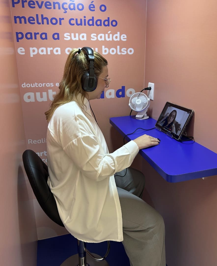 Close-up de uma mulher em uma cabine de atendimento, sentada de perfil em uma cadeira preta, usando fones de ouvido e olhando para um tablet sobre uma pequena mesa azul. O texto promocional na parede rosa diz: "Prevenção é o melhor cuidado para a sua saúde e para o seu bolso".