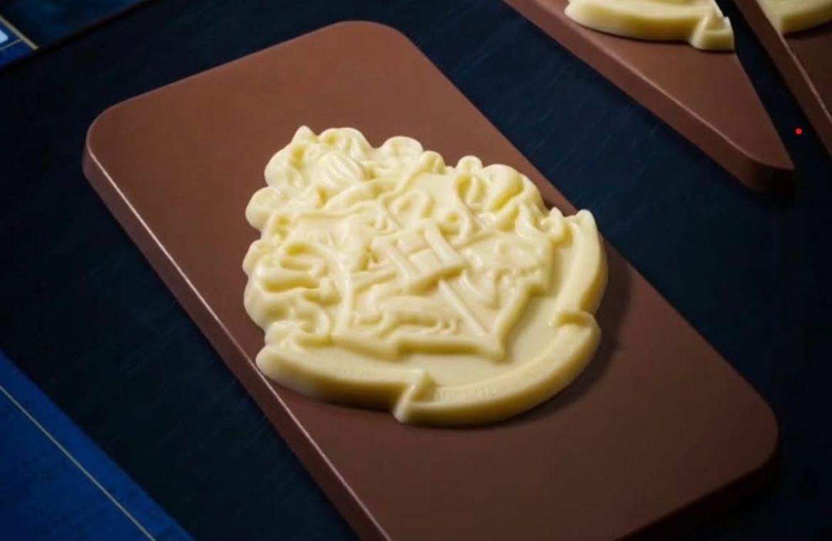 Close-up de um tablete de chocolate ao leite da Cacau Show com o brasão de Hogwarts em relevo feito de chocolate branco. O tablete está apoiado sobre uma superfície escura.
