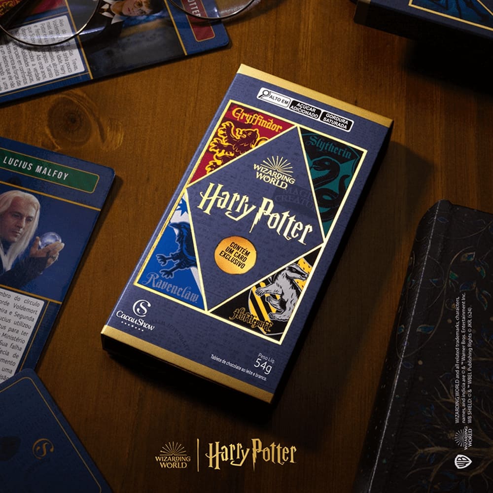 Caixa de chocolate Cacau Show Harry Potter em destaque, com as cores e emblemas das quatro casas de Hogwarts (Grifinória, Sonserina, Lufa-Lufa e Corvinal). A embalagem azul escura indica que contém um "Card Exclusivo". Ao redor, cartas colecionáveis do universo Harry Potter, incluindo uma com a imagem de Lucius Malfoy.